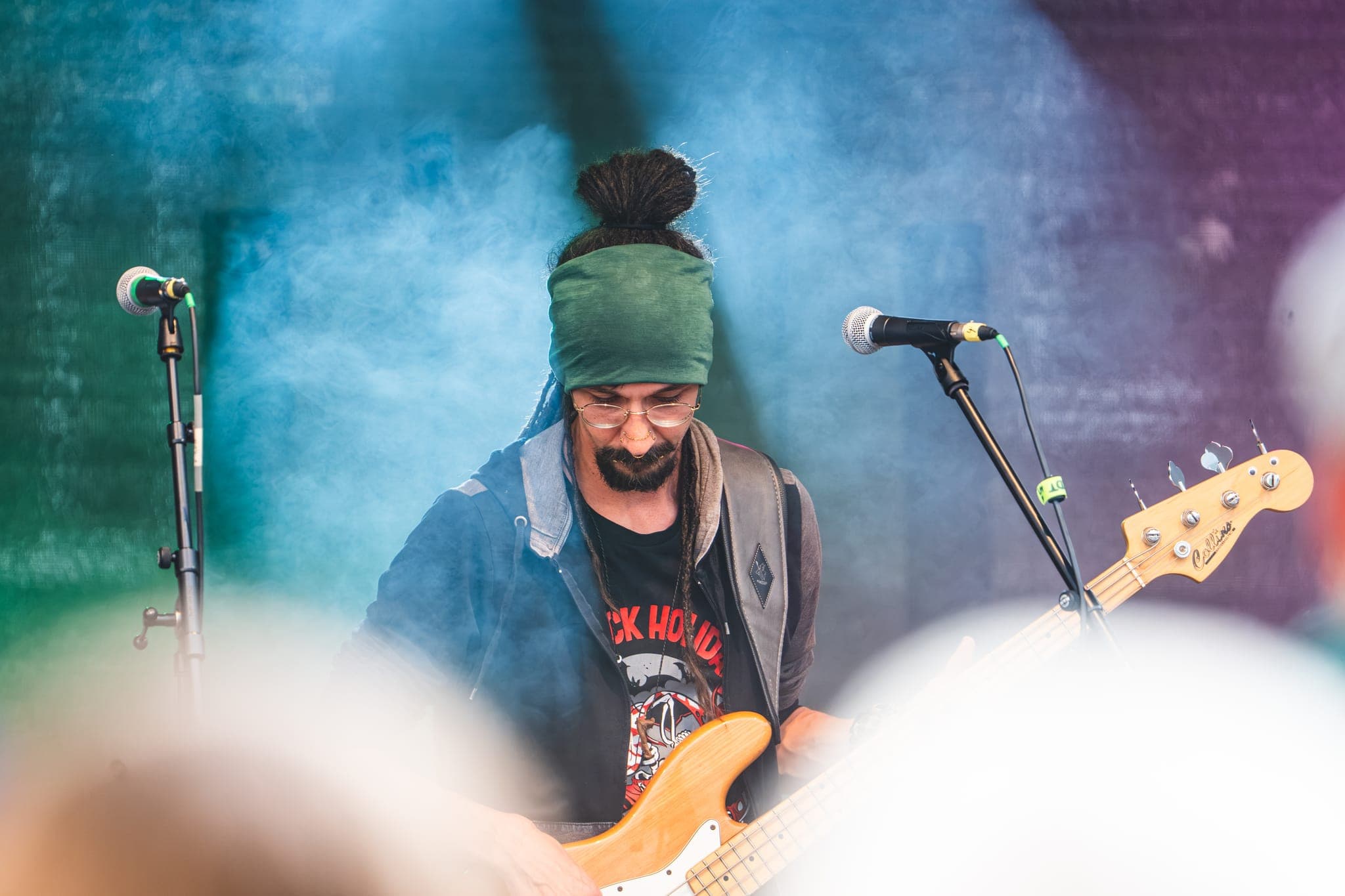 Bassist mit grünem Stirnband und Dreadlocks im Bühnenrauch, durch das Publikum fotografiert